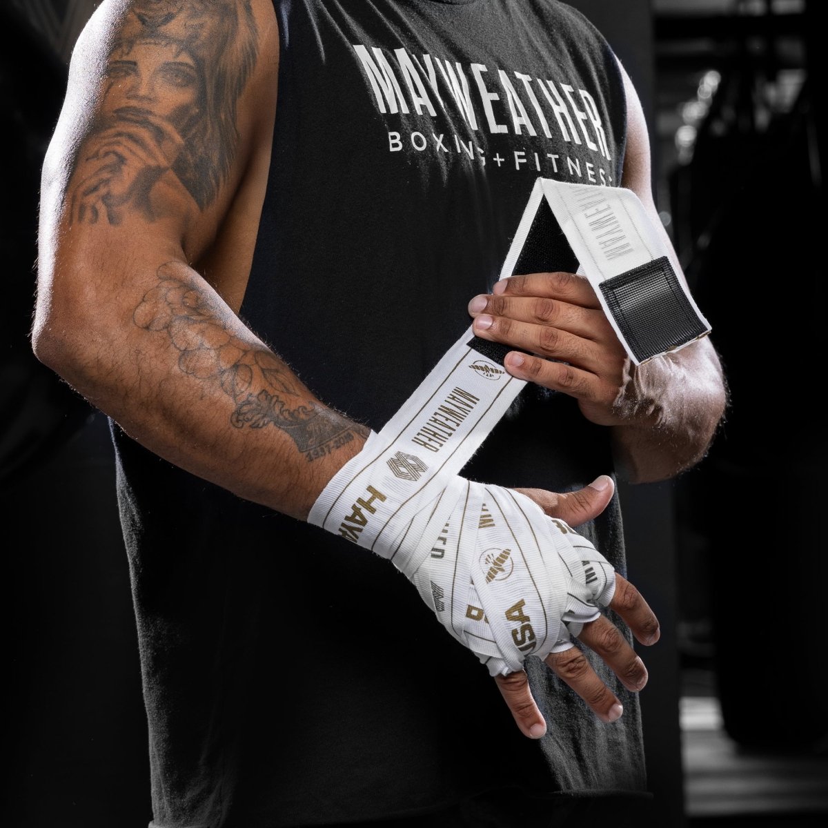 Vista frontal de la mano de un luchador con un vendaje completo usando el vendaje elástico blanco y dorado de 180 pulgadas de Mayweather Hayabusa, con el logotipo de 'MAYWEATHER' visible en el área de la muñeca.